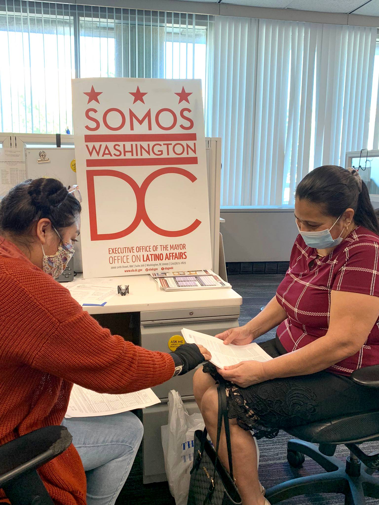 Photo of two people wearing masks, looking at paperwork together. A poster that says "Somos Washington DC" is propped on a table behind them.