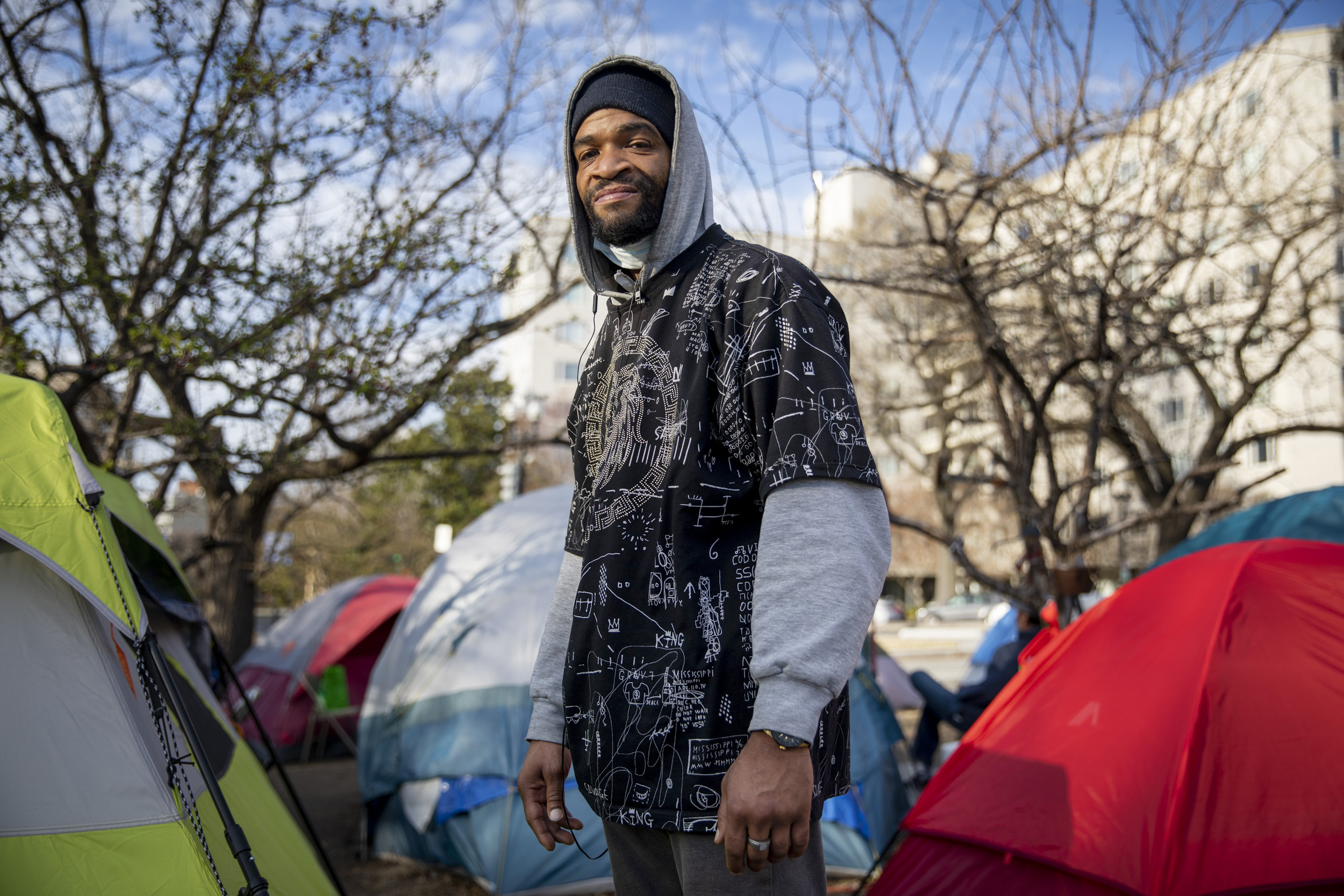 Photo showing a man standing among many tents and several trees, with a road and large building visible behind him.