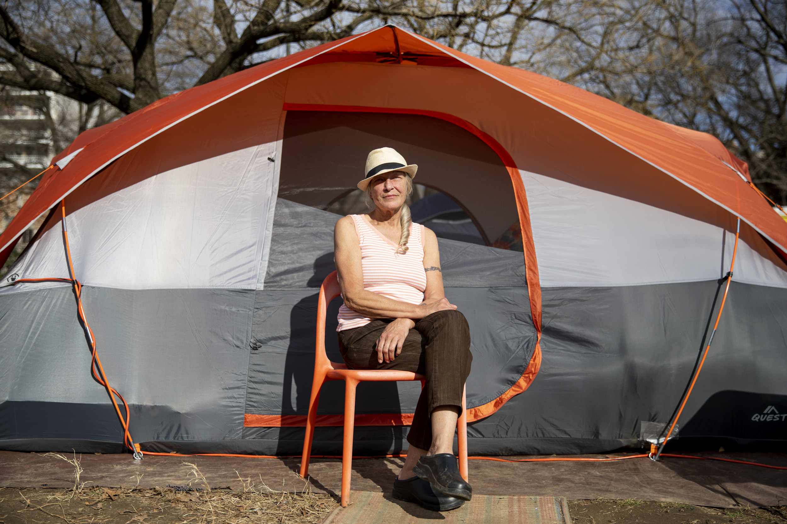 Photo of a woman with gray hair wearing a hat, sitting in a chair centered in front of a large tent.