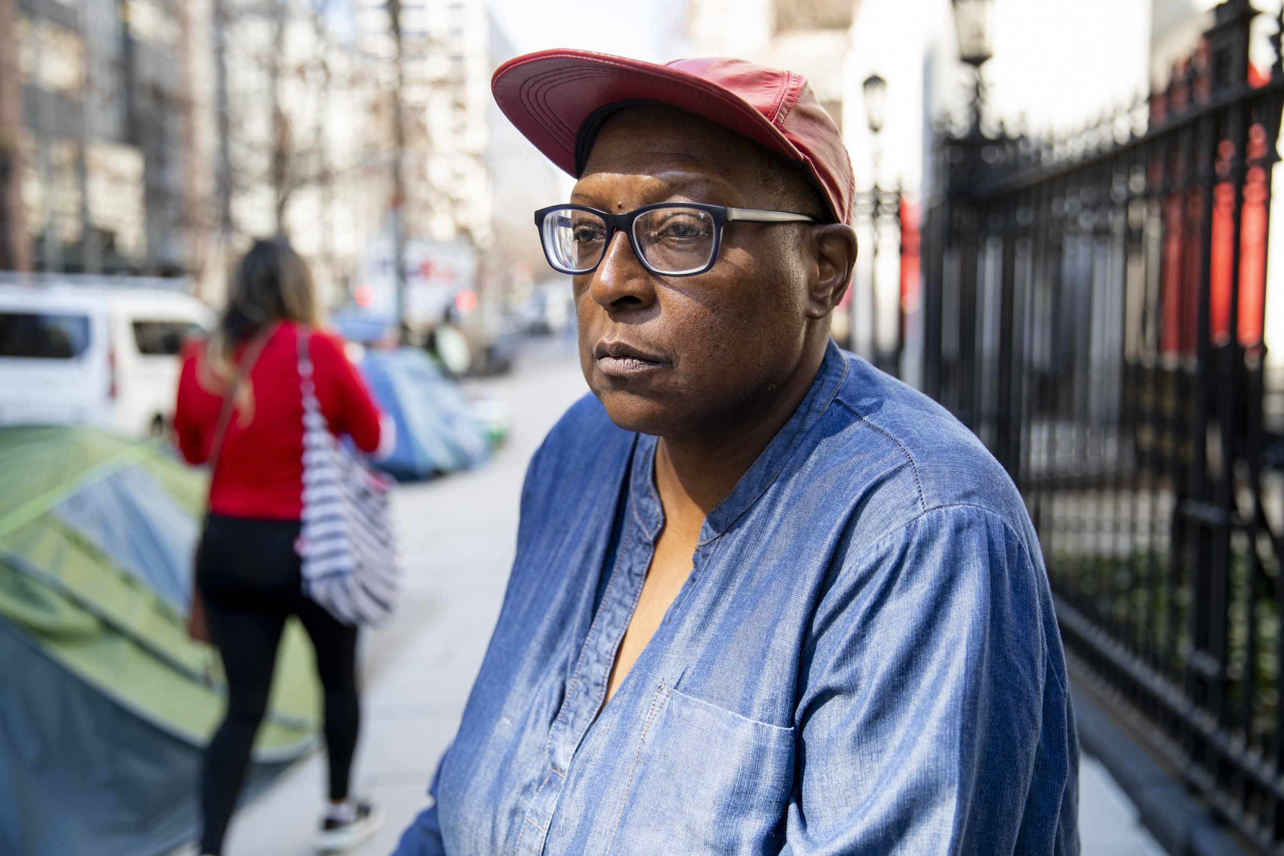 Photo of a woman with a red hat and glasses standing on a sidewalk in DC. A pedestrian, two tents, and a church courtyard are visible behind her.