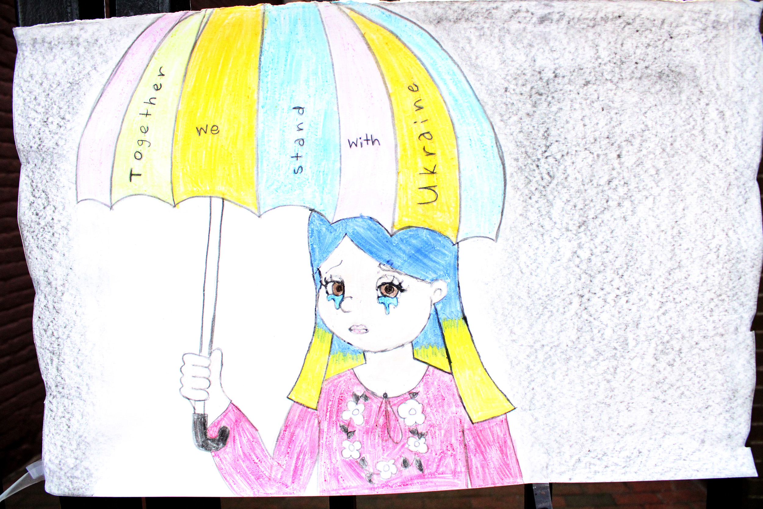 Photo of an illustrated poster featuring a girl holding an umbrella over her head. the colors are most the blue and yellow of the Ukrainian flag and the words "together we stand with Ukraine" are written on the umbrella.