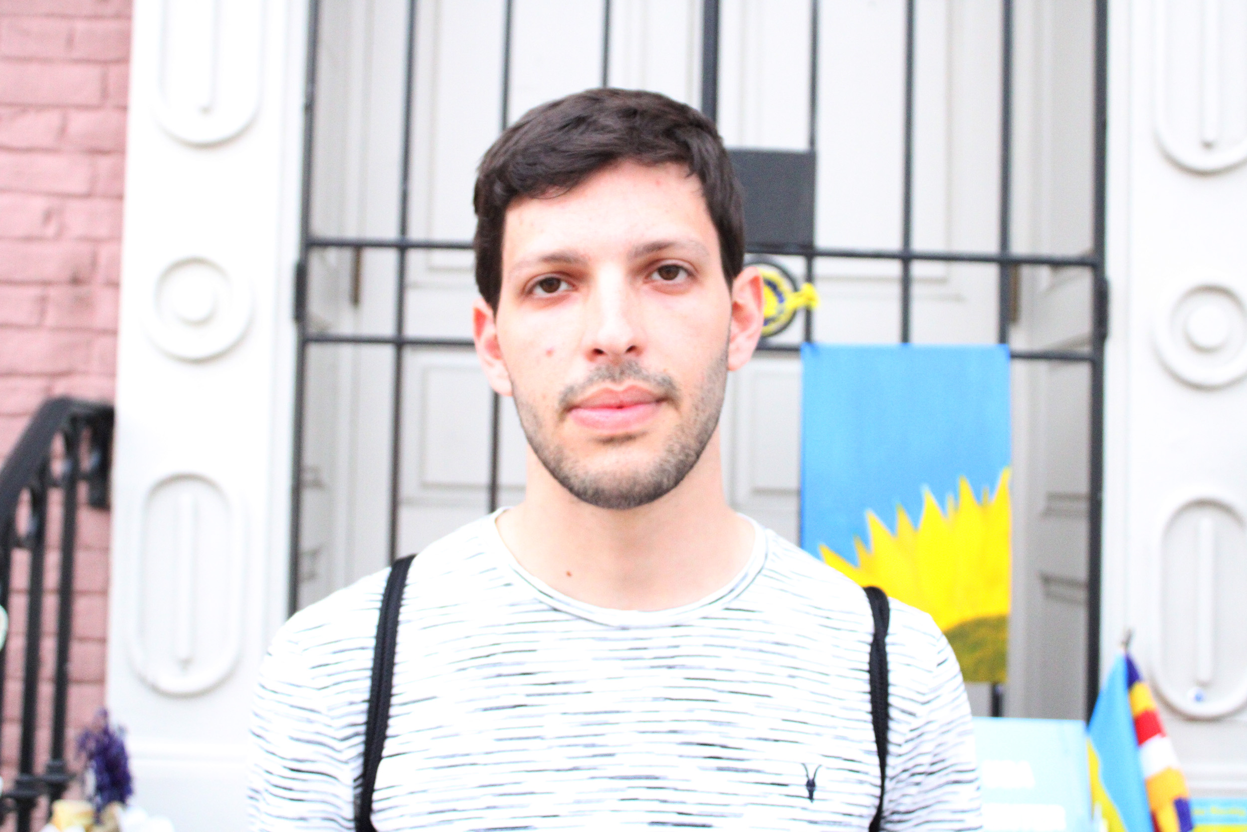 Photo of a man wearing a t-shirt and a string backpack. A painting of a sunflower is visible against a door and brick building behind him.
