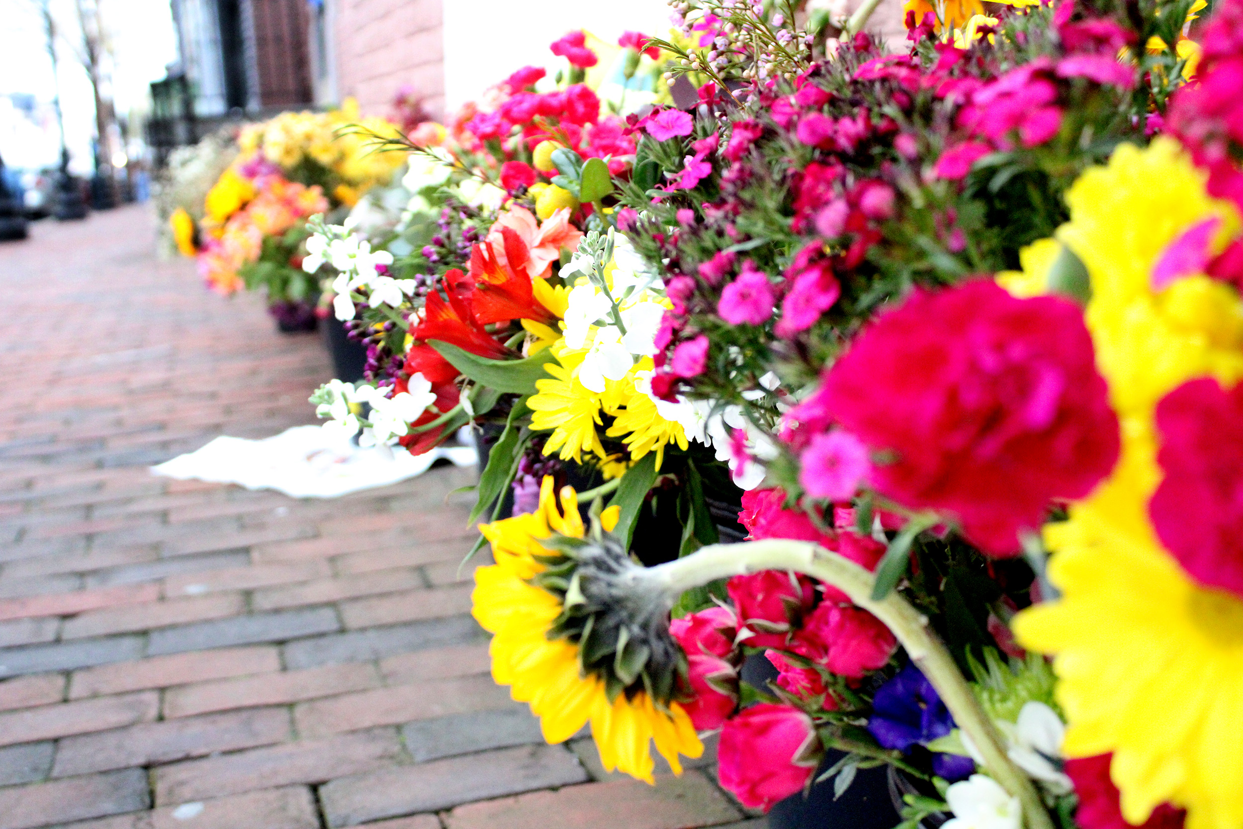 Photo of many brightly colored flowers placed alongside a the Embassy of Ukriane building on a brick sidewalk.