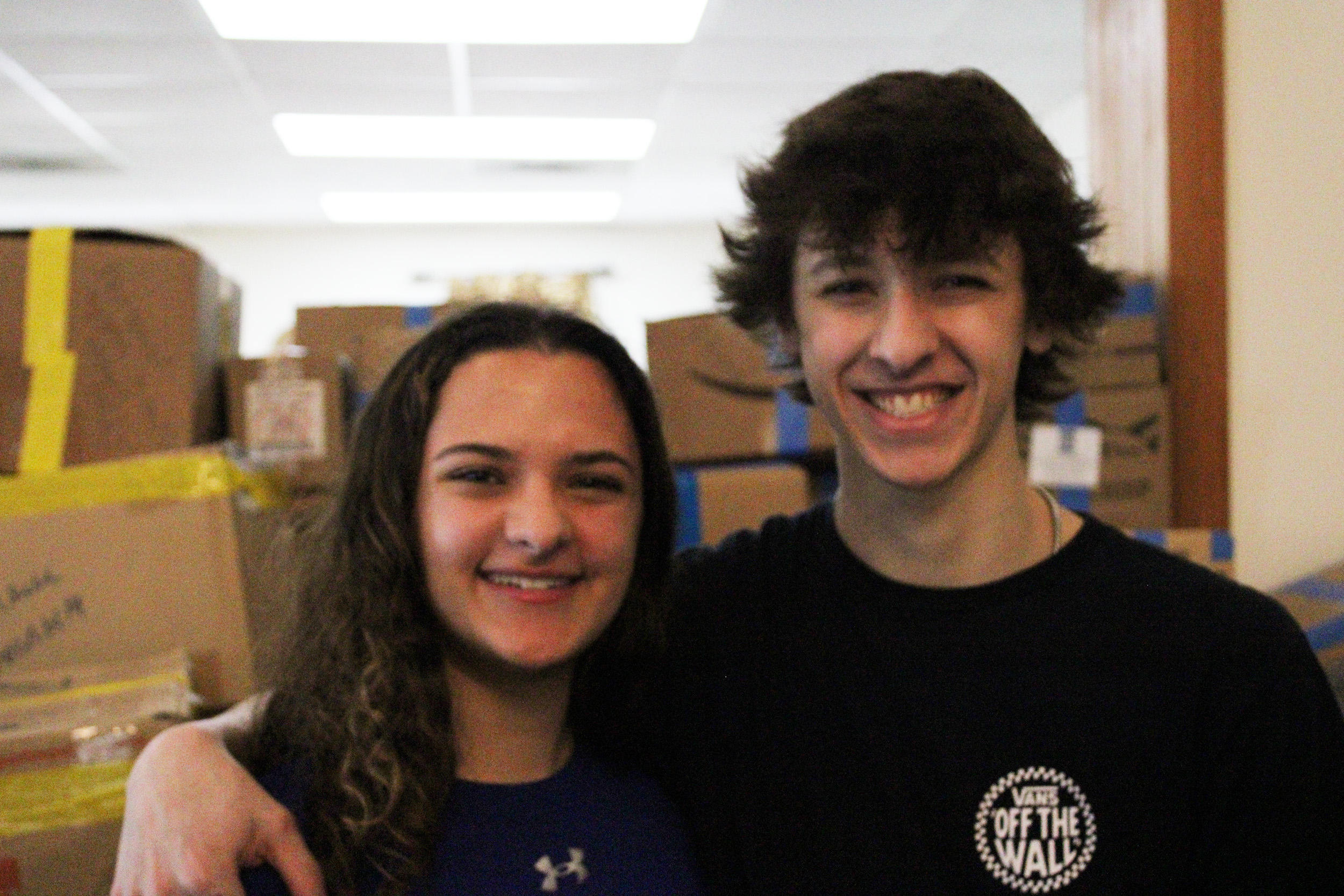 Photo of an adolecent girl and boy with their arms around each other's shoulders. Many boxes of donatoins for Ukraine can be seen stacked behind them.