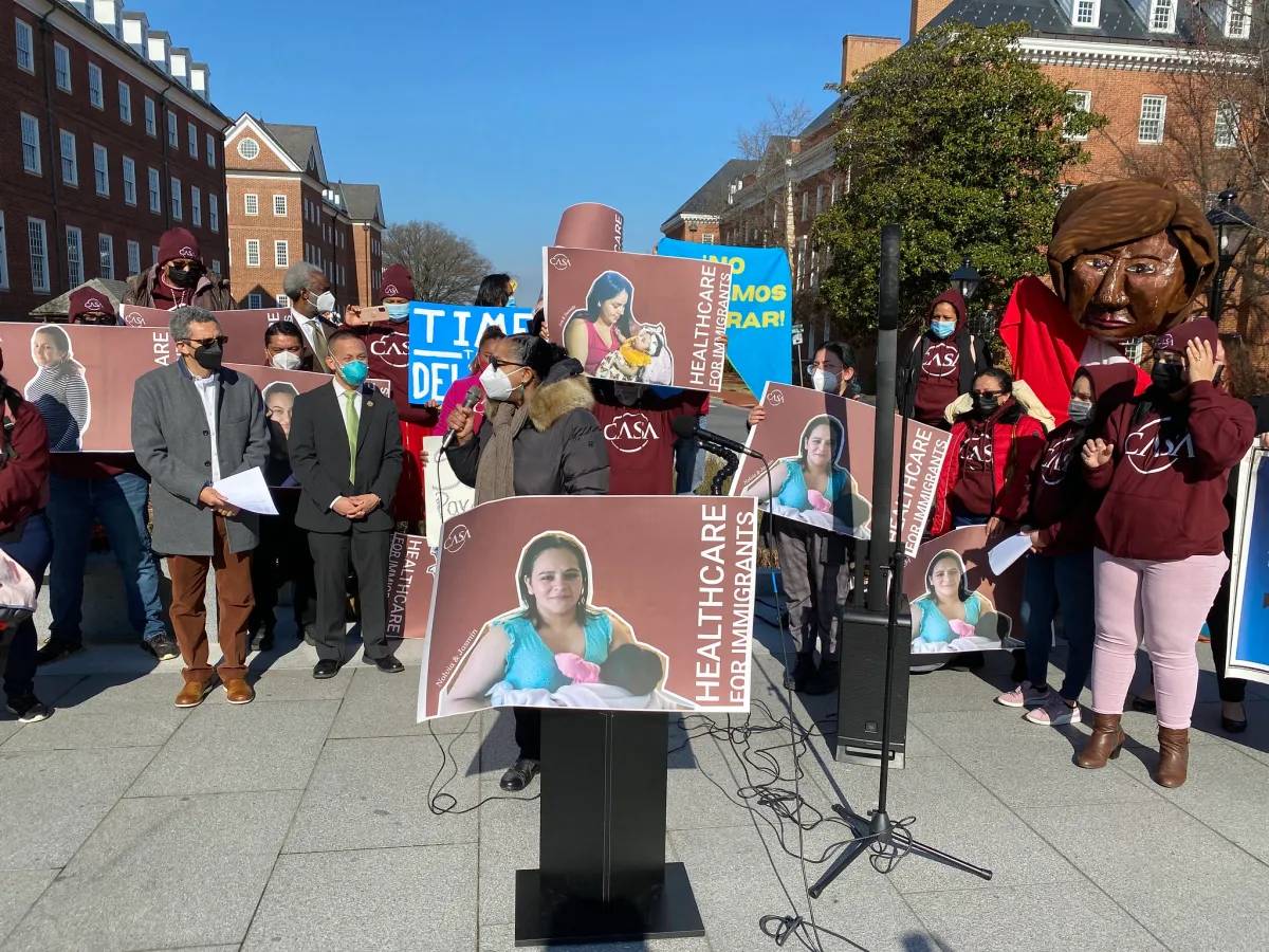 photo of a woman standing at a podium bearing a "health care for immigrants" sign