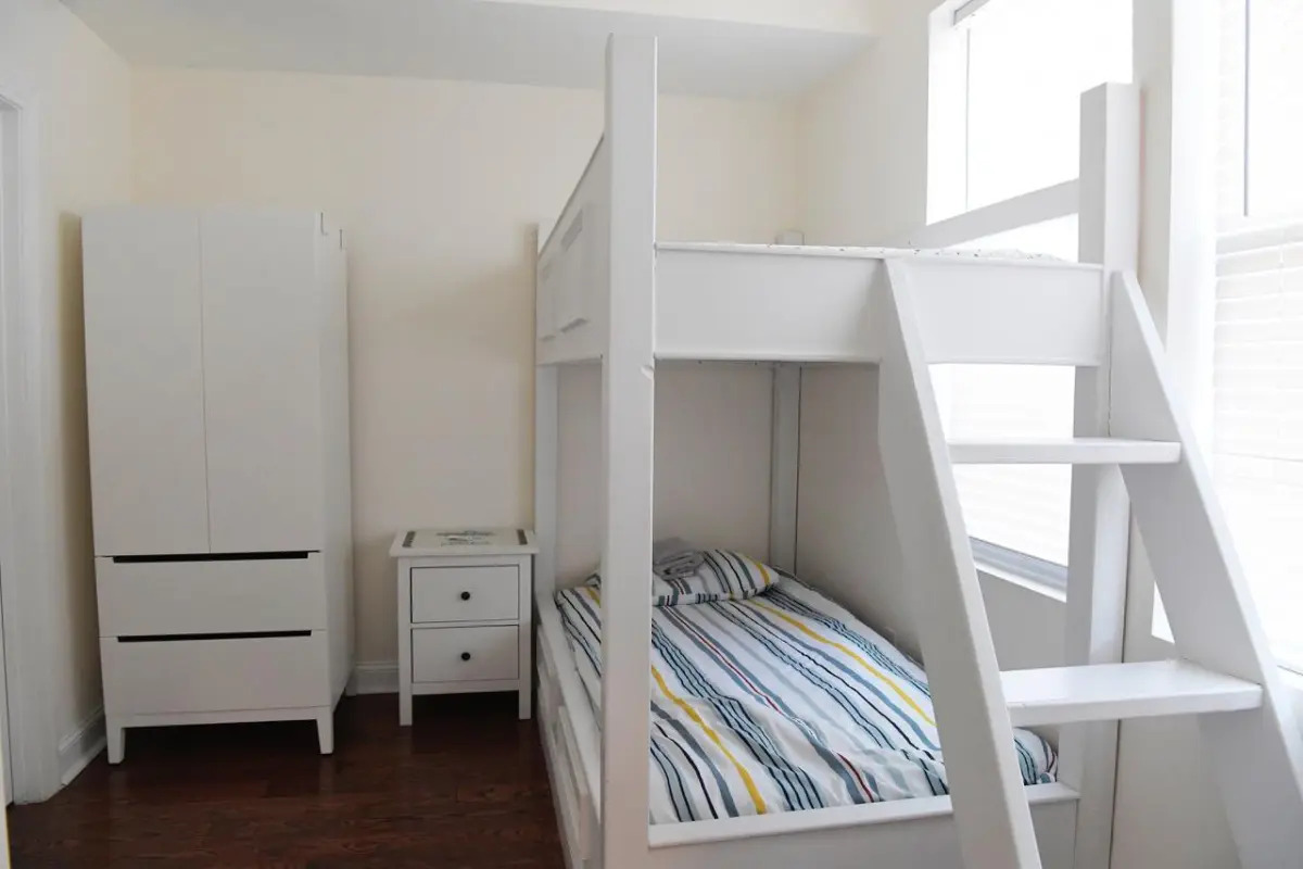 Photo of a set of bunk beds in a dormitory at the shelter. They are painted white with birghtly striped sheets on the bottom bunk mattress. A matching nightstand and dresser are scene next to the beds.