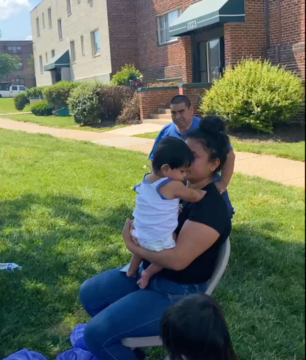 A photo of a very small child being held by a woman sitting in a chair outdoors in the heat. Apartment buildings are visible behind her on the other side of a walking path.