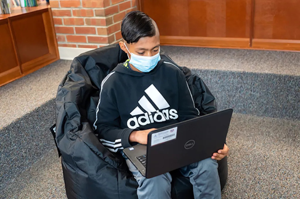A student sitting in a bean bag chair with a laptop, wearing a face mask to protect against COVID-19