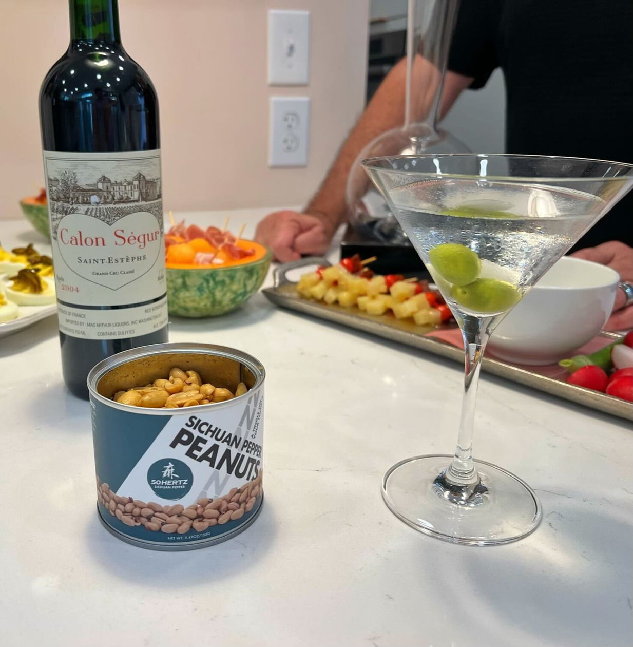 A photo of a can of Sichuan Pepper Peanuts on a counter next to a bottle of wine, a martini, and platters of food.