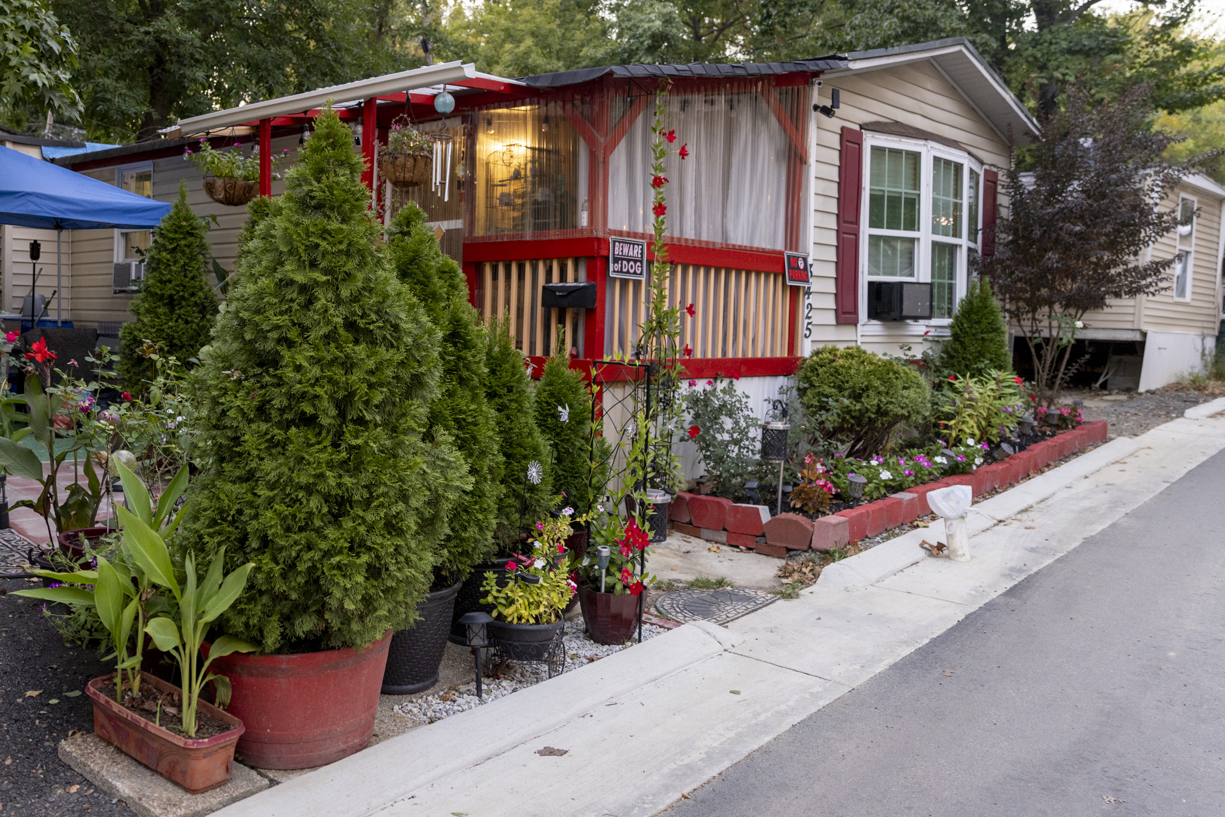 Photo showing a mobile home with a porch attaached with bright red trim and a number of potted shrubs and flowers outside next to a flowerbed.