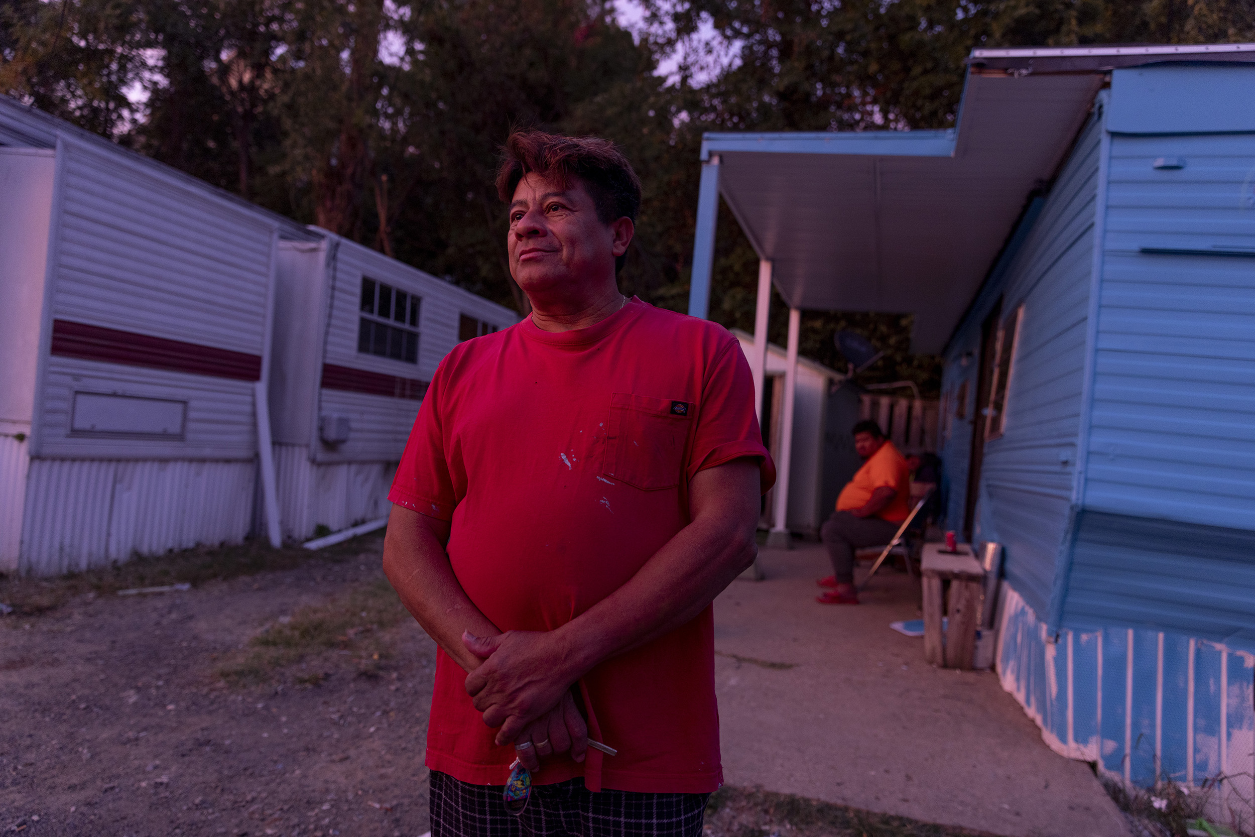Photo of a man standing outside of a manufactured home. Someone else is visible behind him, sitting in a chair under an awning.