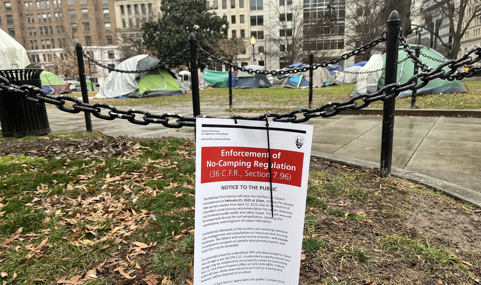 A sign reading "Enforcement of no-camping regulation" is attached to a chain at the border of McPherson Square park. Tents are visible behind it.