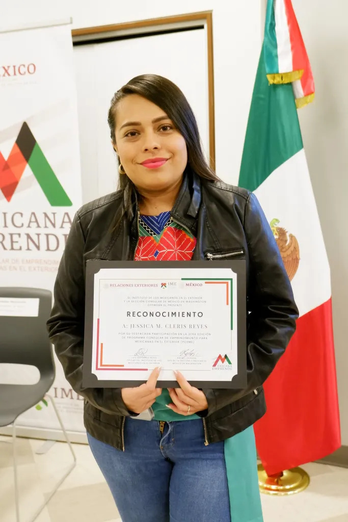 Reyes poses with her certificate, next to a Mexican flag and in front of signage for the Mexicana Emprende program.