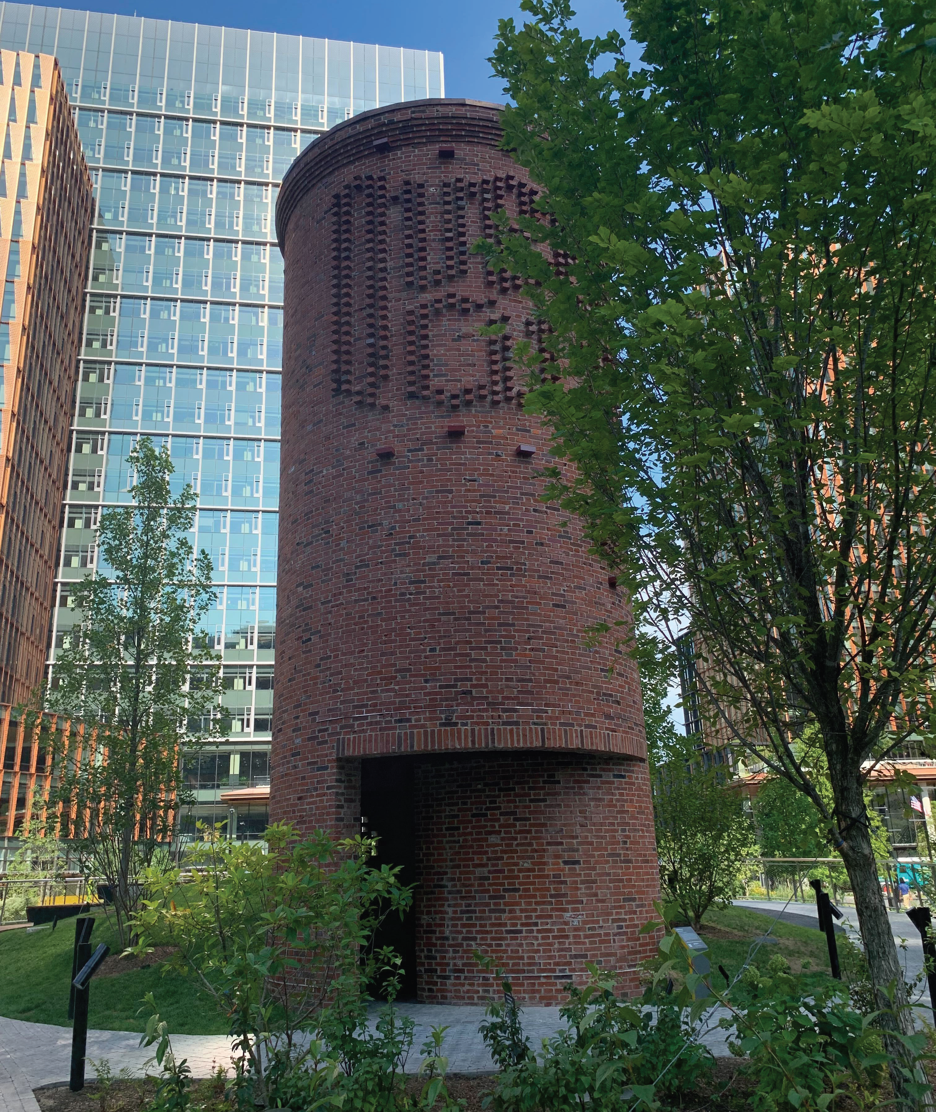 Photo of a red brick tower with an entrance, surrounded by small trees and tall buildings.