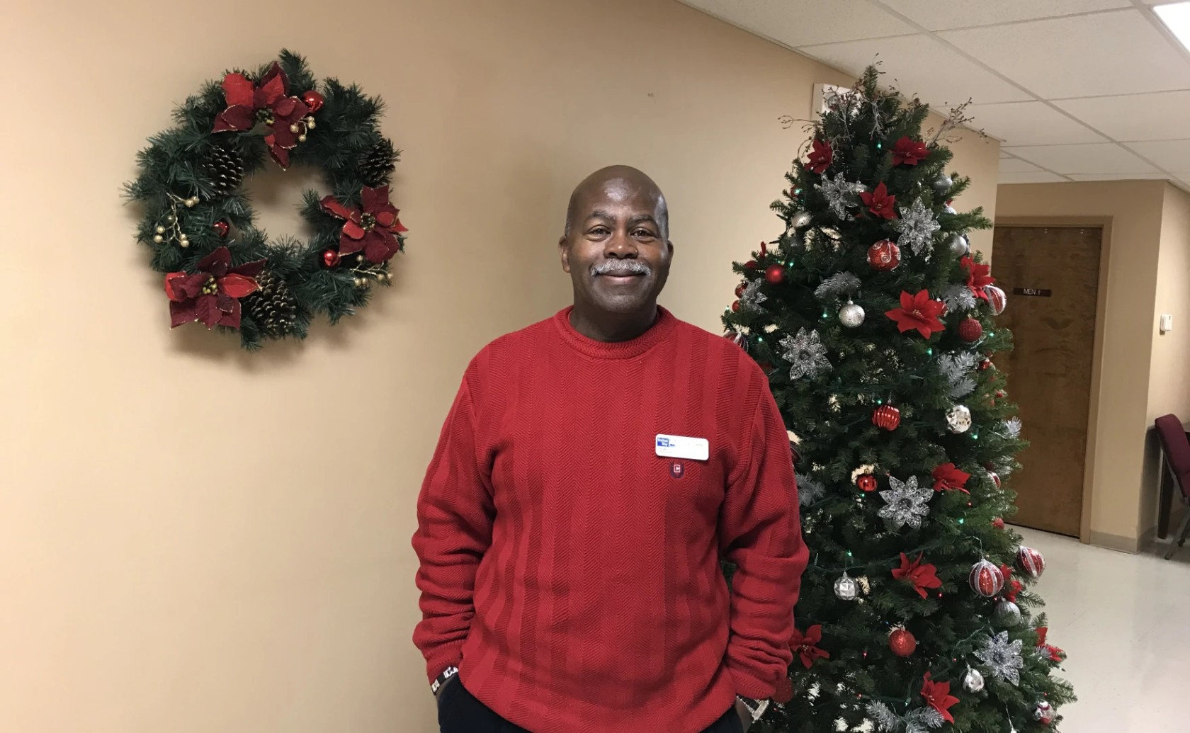 Photo of a man standing between a Christmas tree and a Christmas wreath.