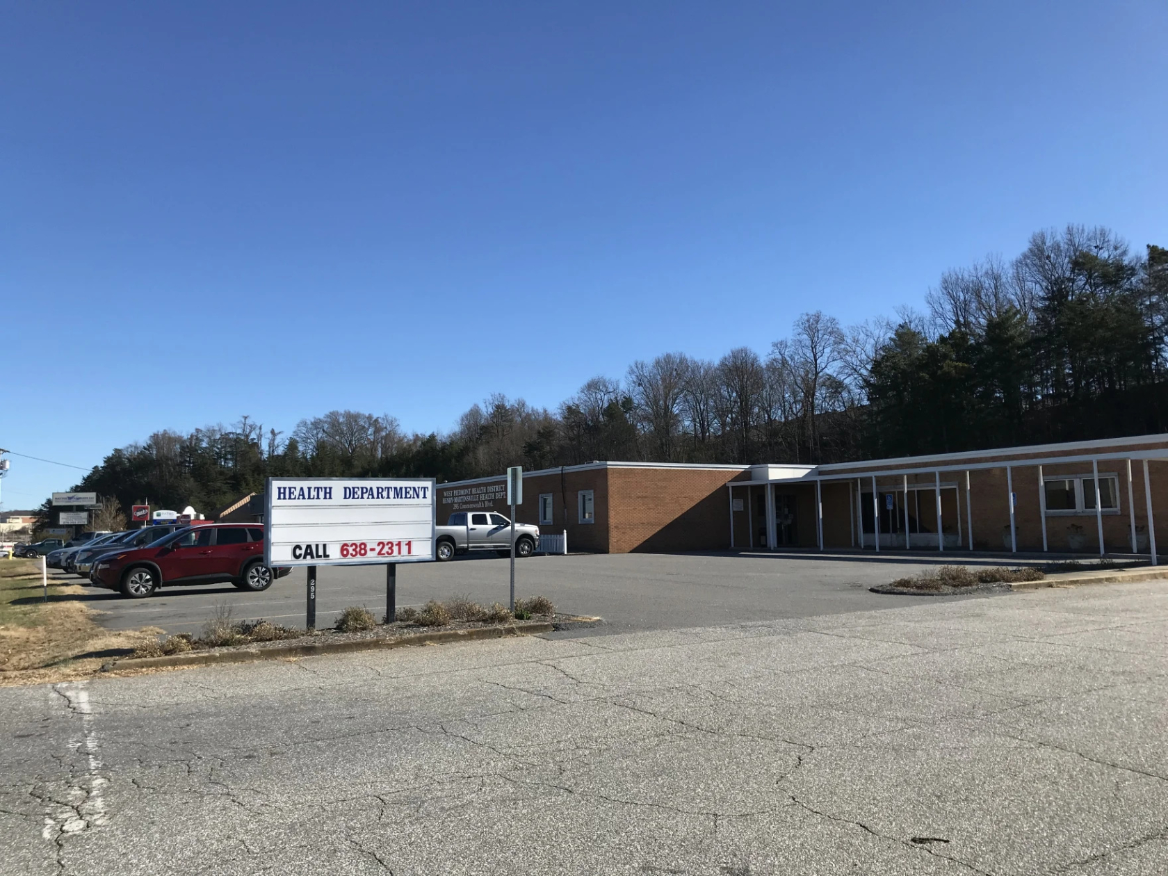 Photo showing a one-story brick building and adjacent parking lot. The building says "West Piedmont Health District, Henry-Martinsville Health Dept." The sign in the parking lot says "Health Department Call 638-2311".