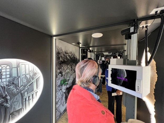 A teacher wearing headphones and looking at a screen. Behind them, two scenes can also be seen on the wall of the trailer, one that appears to be an historic photo and another that appears to be a modern mural.