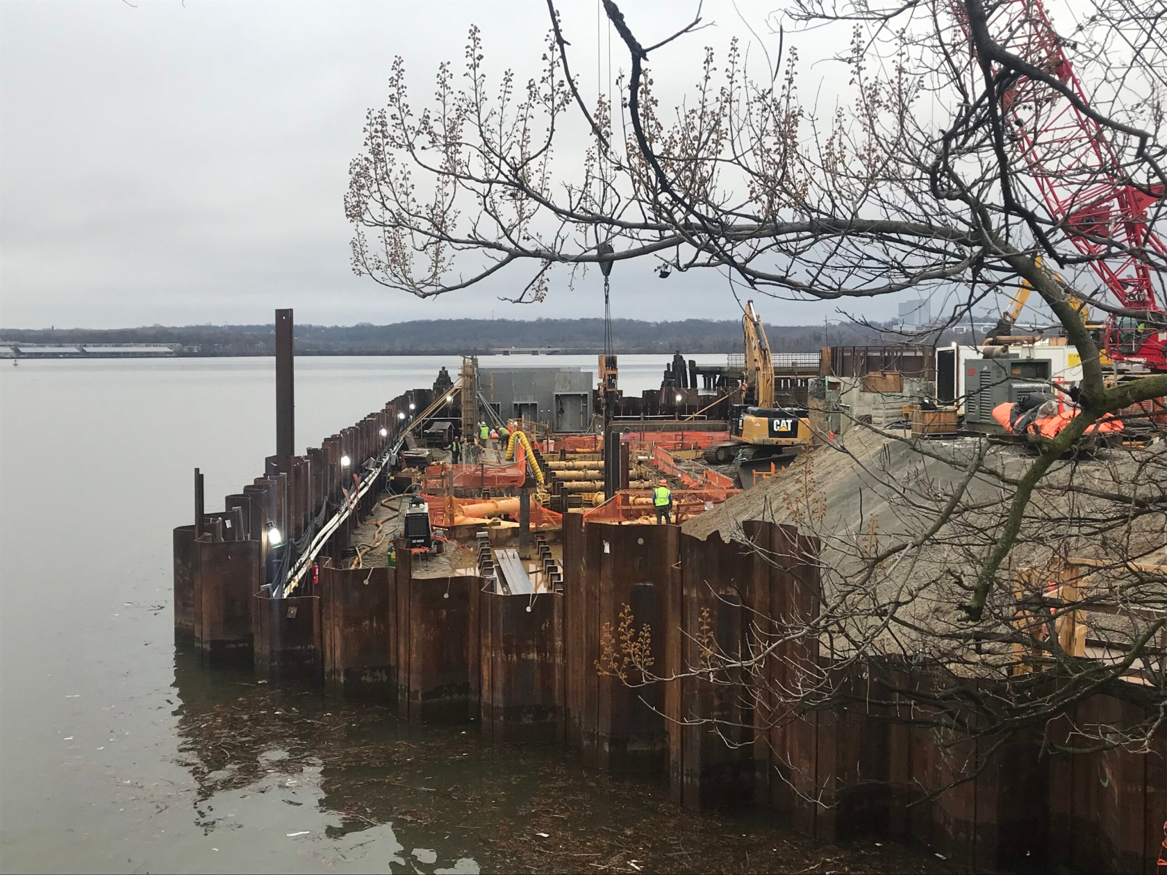Photo showing construction at the edge of the Potomac River. A tree stretches across the foreground and the opposite riverbank is visible in the background.
