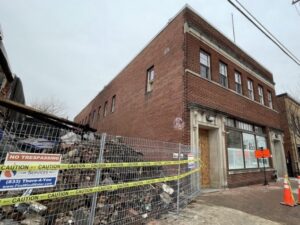Photo showing rubbel behind wire fenscing and caution tape, next to a brick building with boarded up doors and windows.